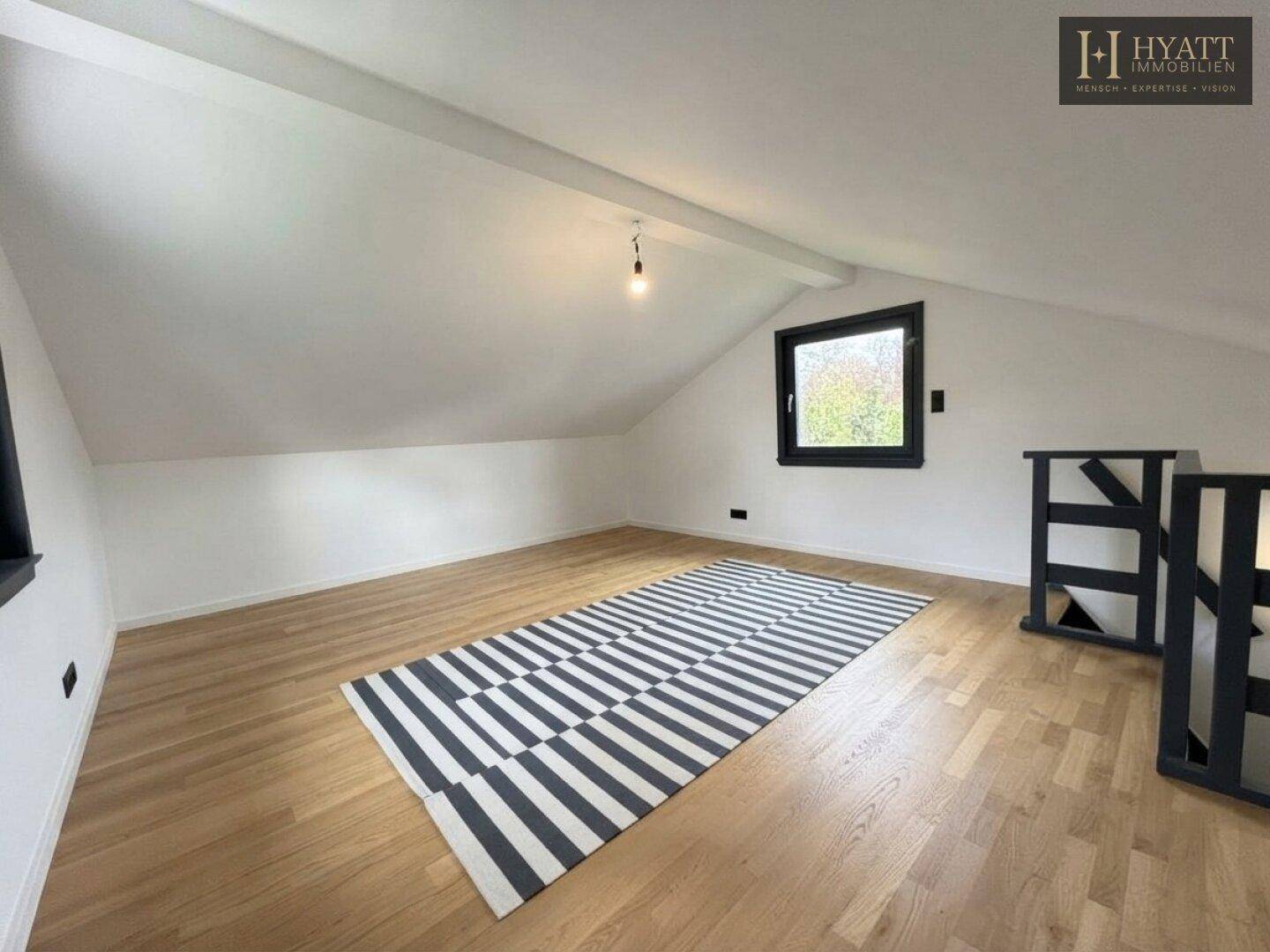 Geräumiges Dachzimmer mit Holzboden, schrägen Wänden und einem modernen schwarz-weiß gestreiften Teppich.