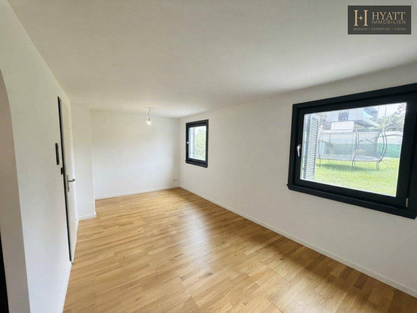 Heller Raum mit Holzboden und großen Fenstern, die viel Tageslicht hereinlassen und Gartenblick bieten.