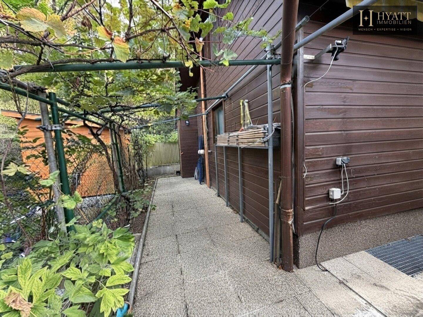 Seitenansicht des Holzhauses mit einem schmalen gepflasterten Weg und Rankpflanzen.
