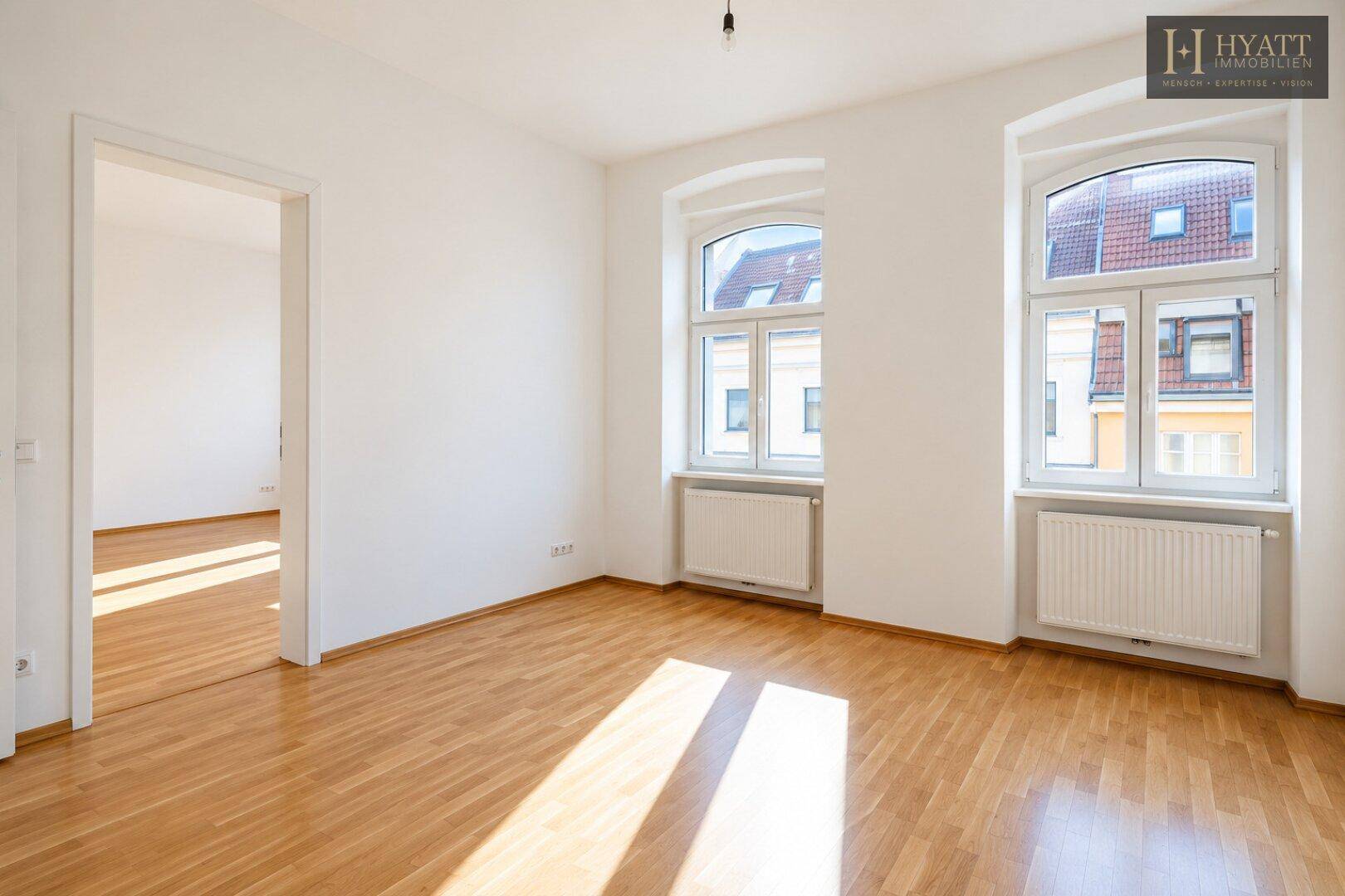 Helles Zimmer mit Parkettboden, weißen Wänden und zwei großen Fenstern, die viel Tageslicht hereinlassen.