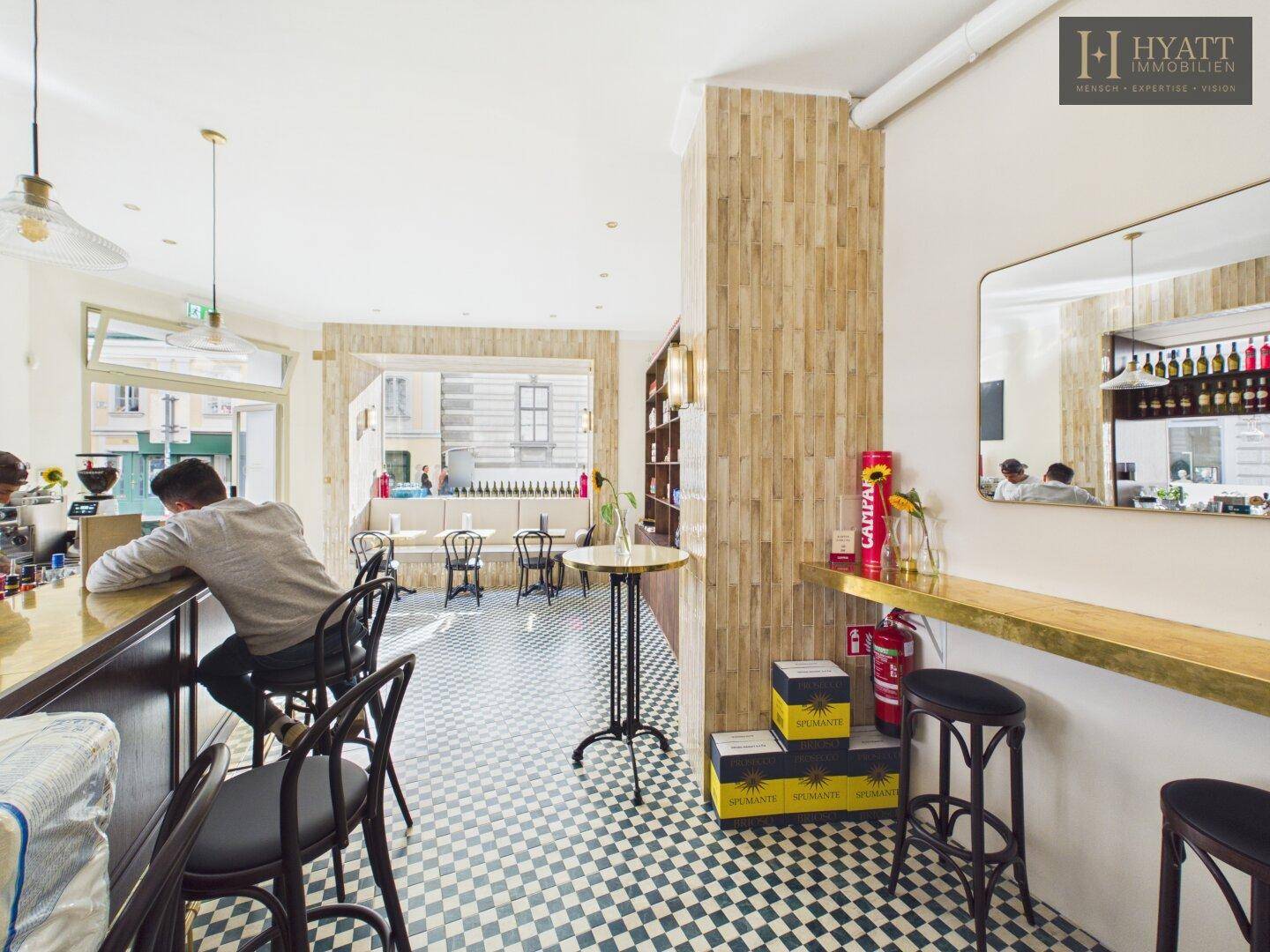 Innenansicht eines Cafés mit Bar, Sitzbereich und Blick auf die Straße durch große Fenster.