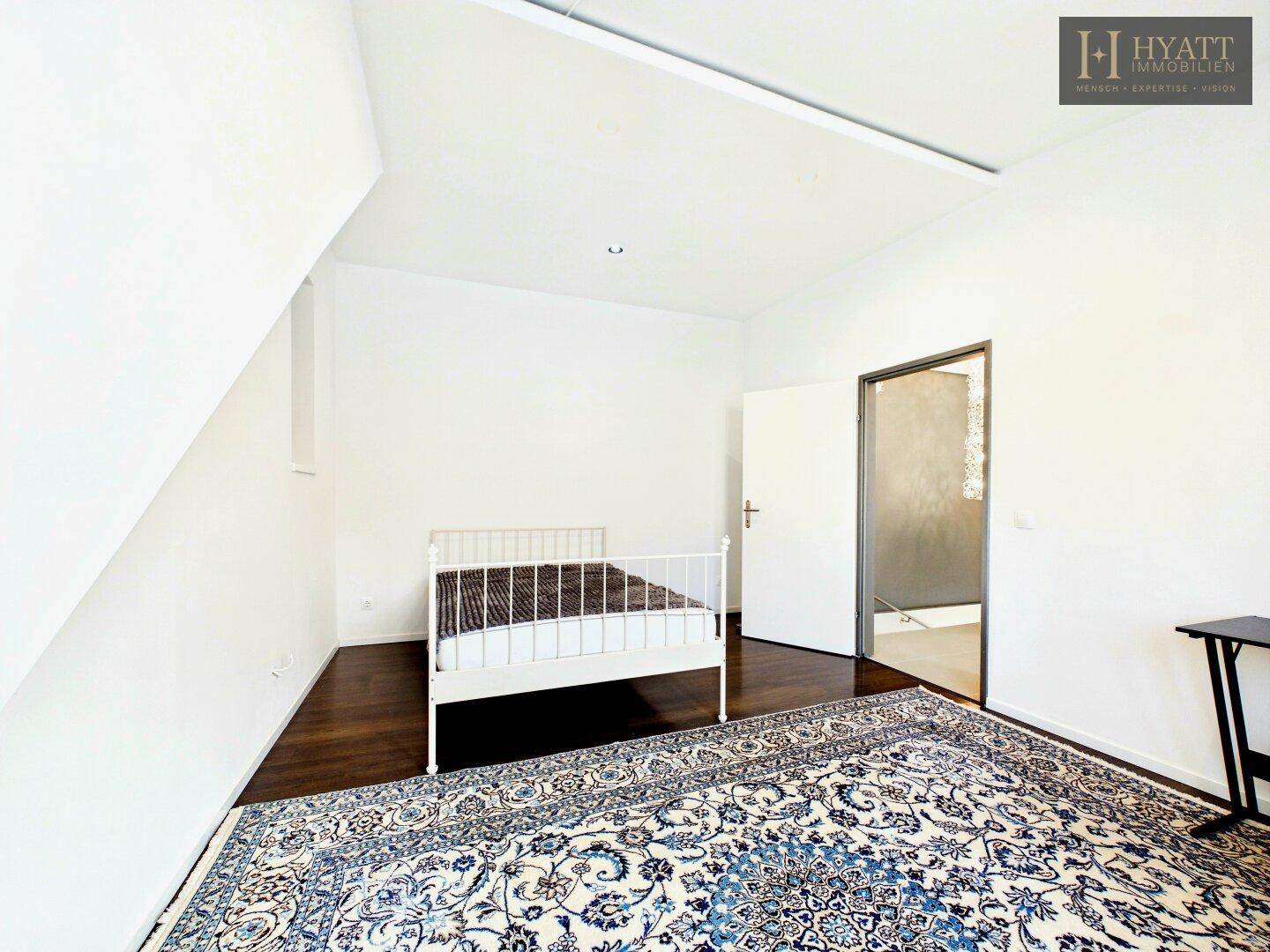 Helles Schlafzimmer im Dachgeschoss mit Holzboden, Teppich und Zugang zu weiteren Räumen.