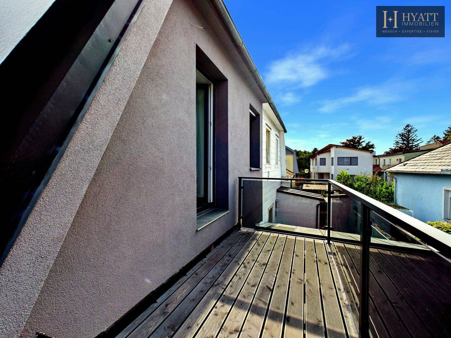 Geräumiger Balkon mit Holzbelag und modernem Glasgeländer an der Hausfassade unter blauem Himmel.