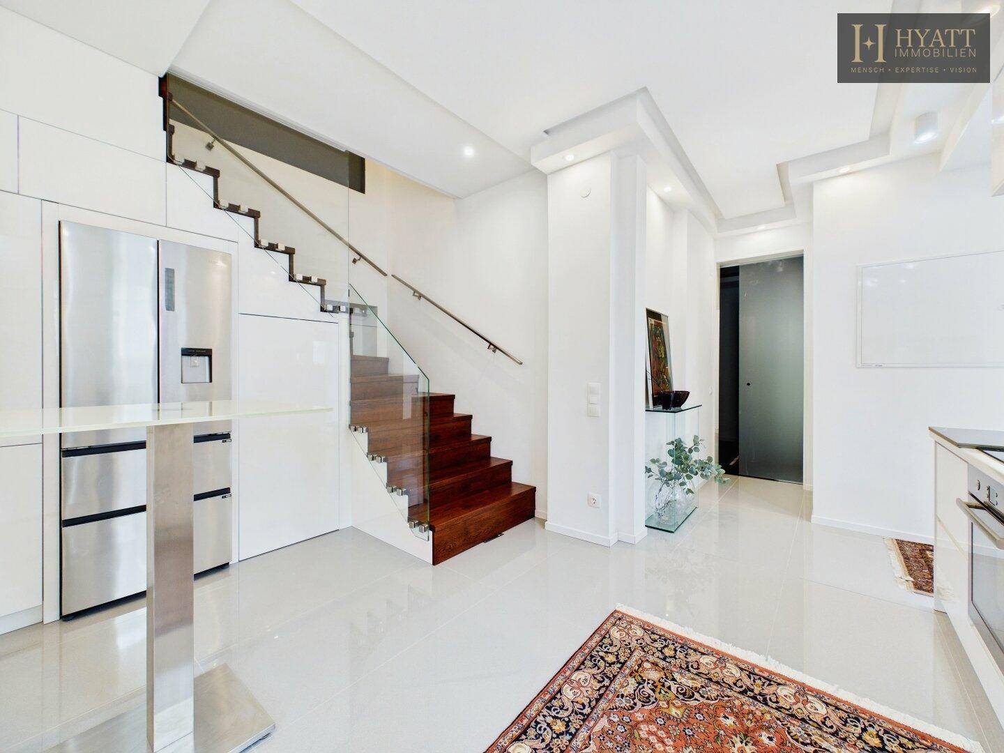 Offene Küche mit modernen Geräten, weißer Einrichtung und eleganter Holztreppe im Hintergrund.