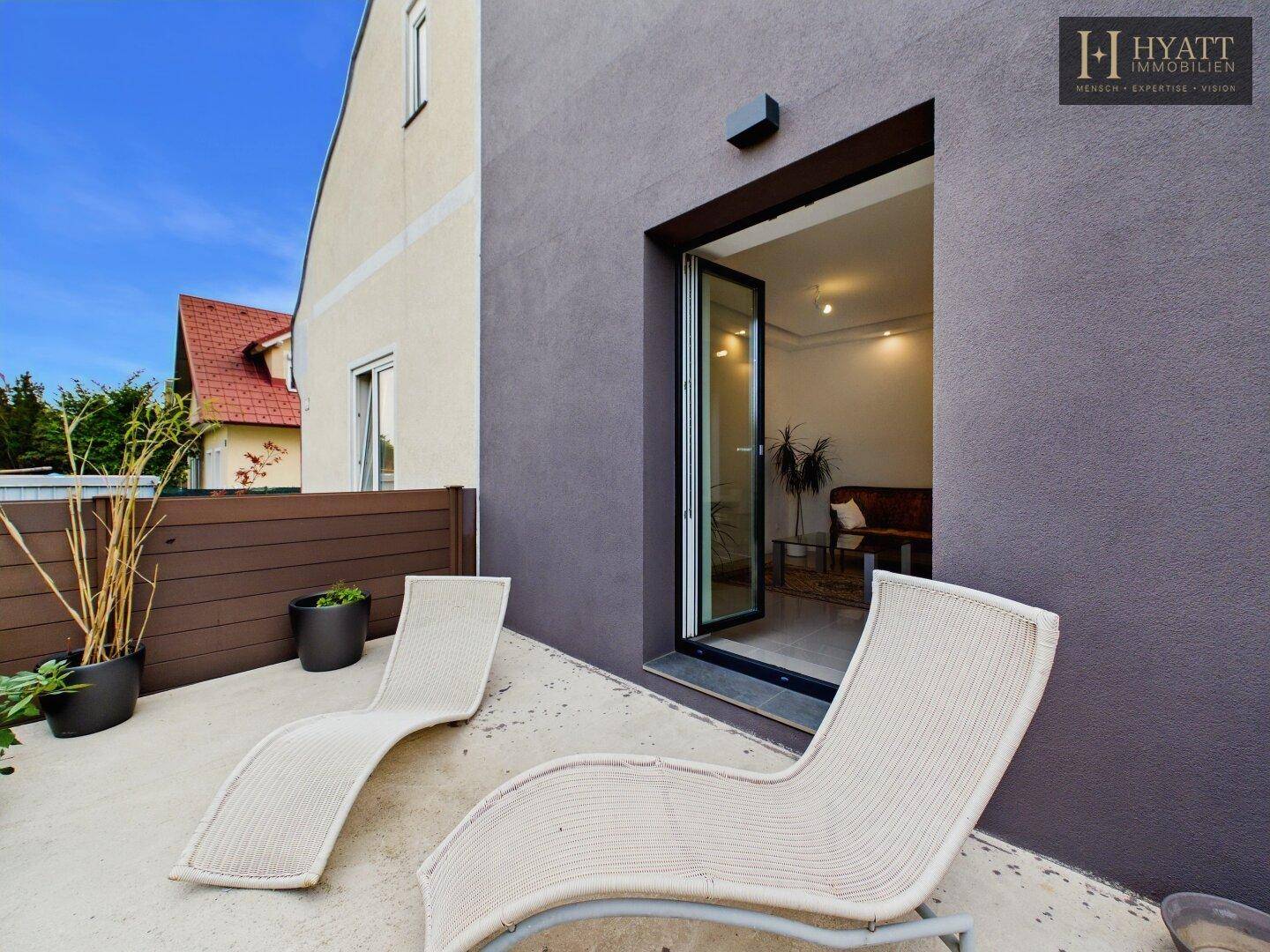 Gemütliche Terrasse mit Liegestühlen und direktem Zugang zu einem hellen Innenraum durch eine Glastür.