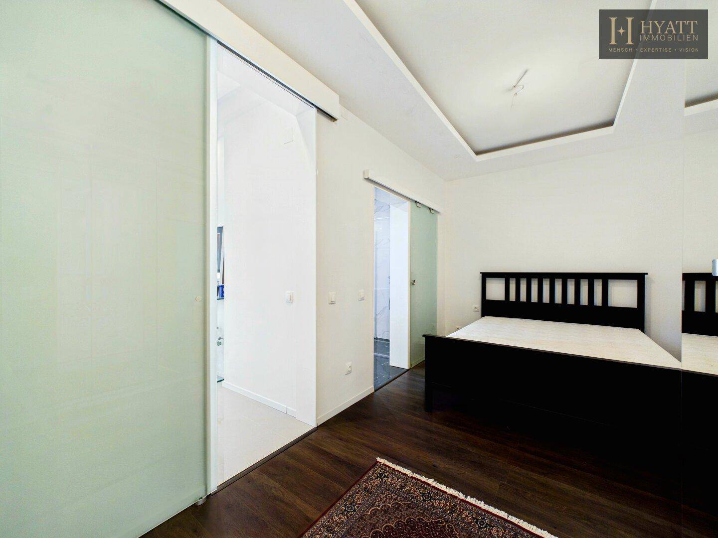 Schlafzimmer mit dunklem Holzboden, minimalistischem Bett und Schiebetüren zu Bad und Ankleide.