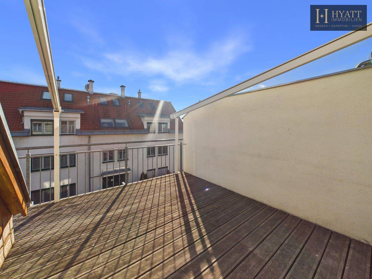 Sonniger Balkon mit Holzdielen und Blick auf die umliegenden Dächer.