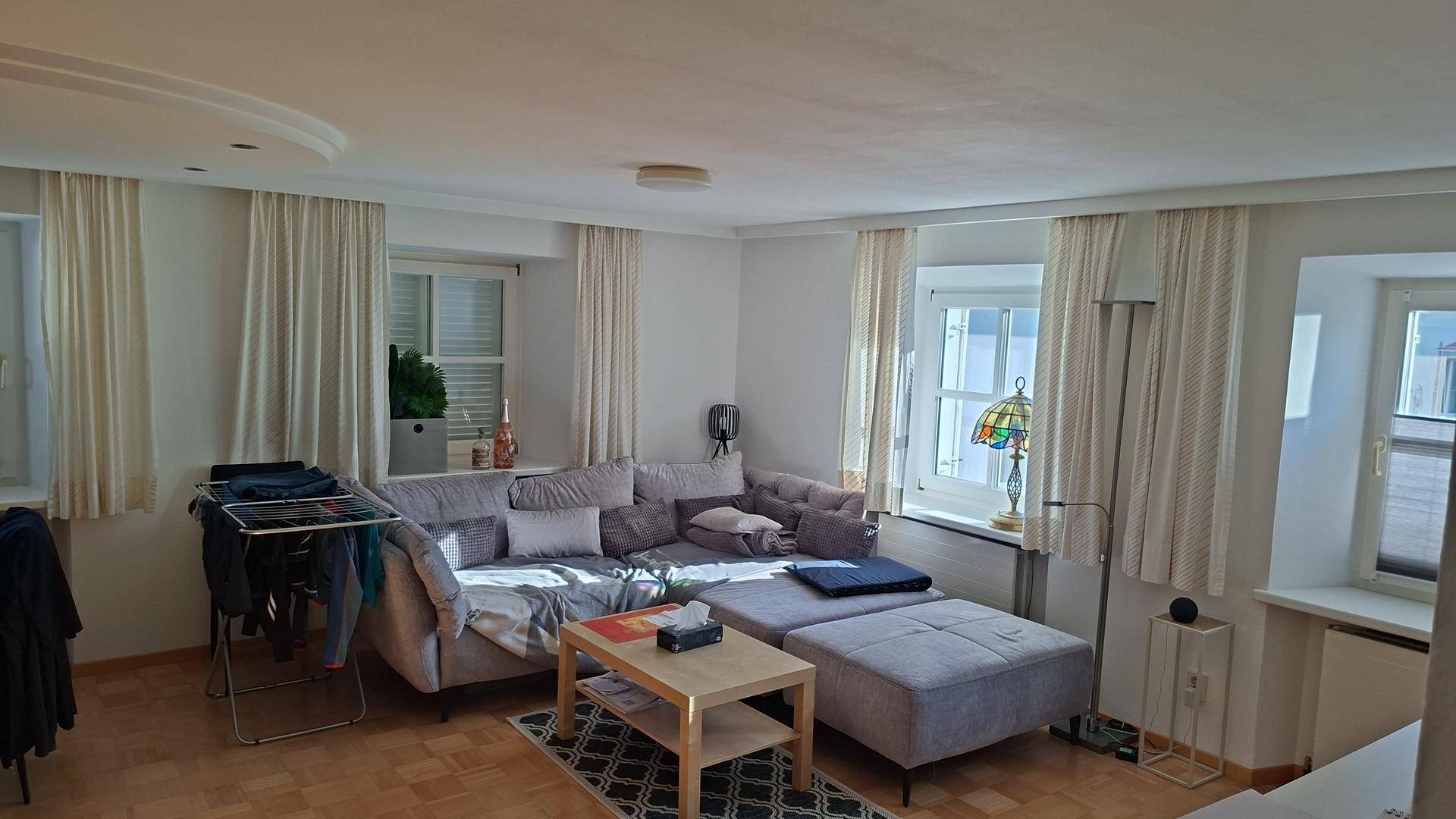 Helles Wohnzimmer mit großem Ecksofa, hellem Parkettboden und mehreren Fenstern mit Vorhängen.