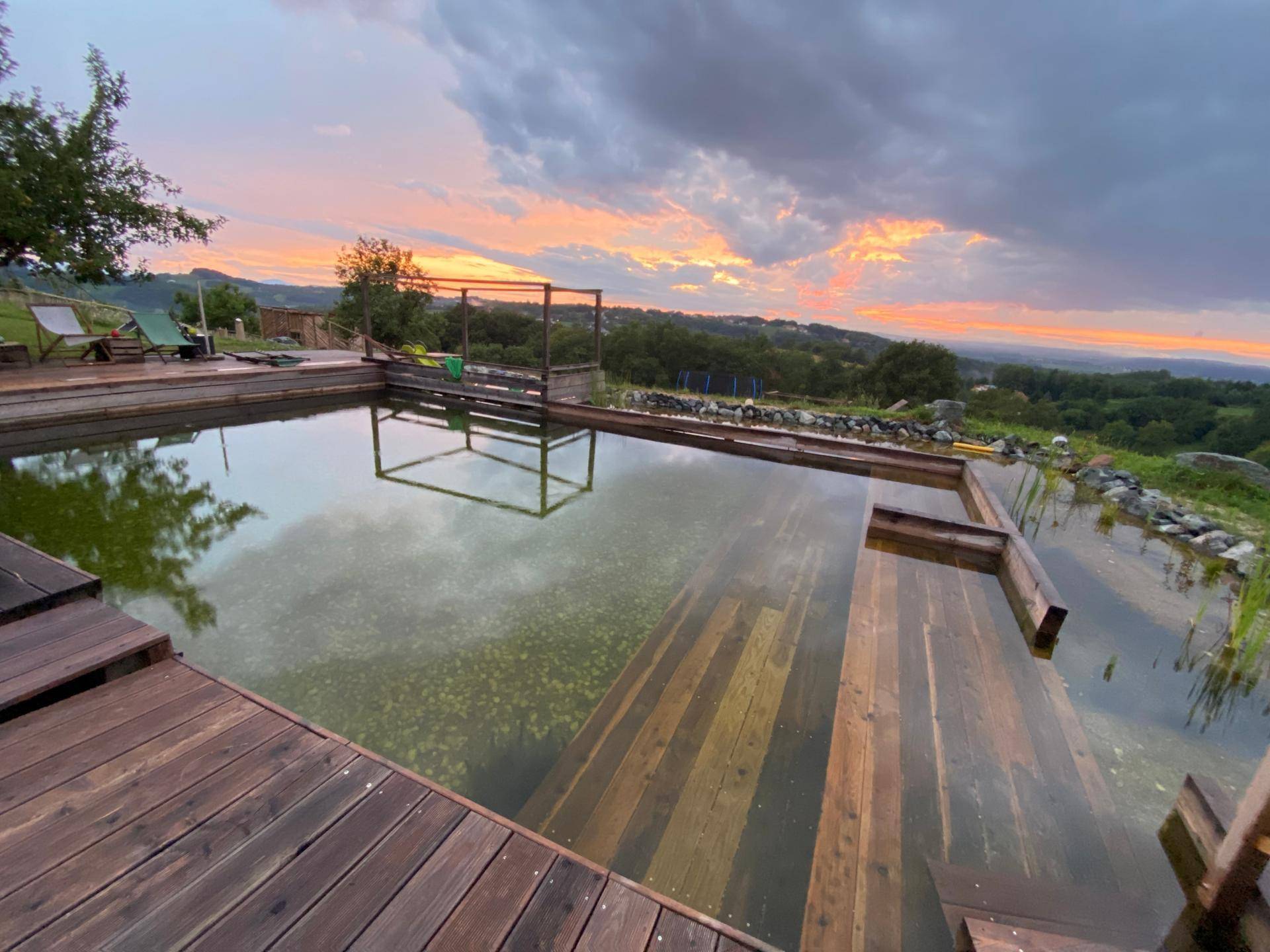 Natürlicher Schwimmteich mit Holzdeck und Blick auf die sanfte Hügellandschaft bei Sonnenuntergang.