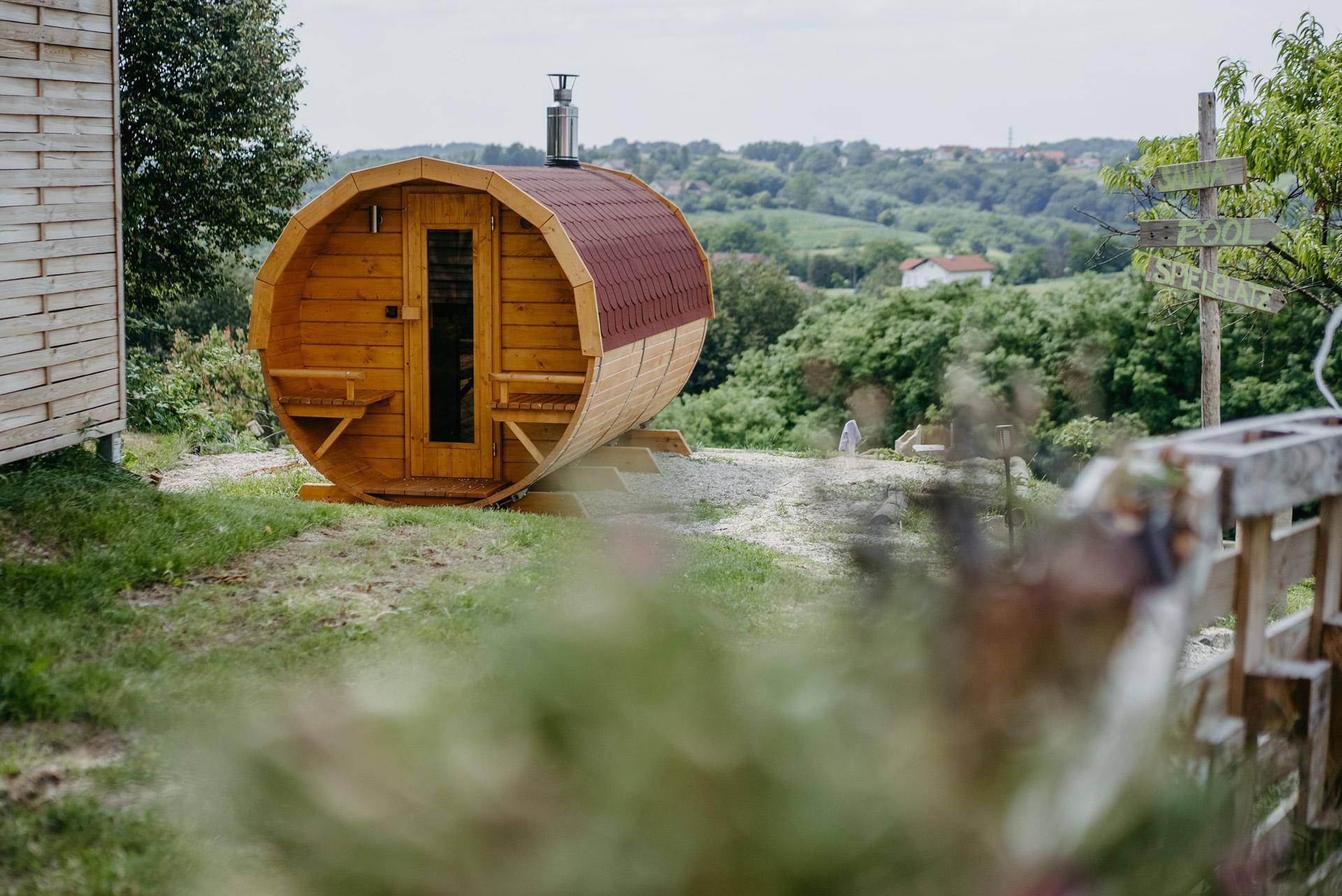Eine rustikale Fasssauna aus Holz steht in einer grünen Hügellandschaft mit weitem Ausblick.