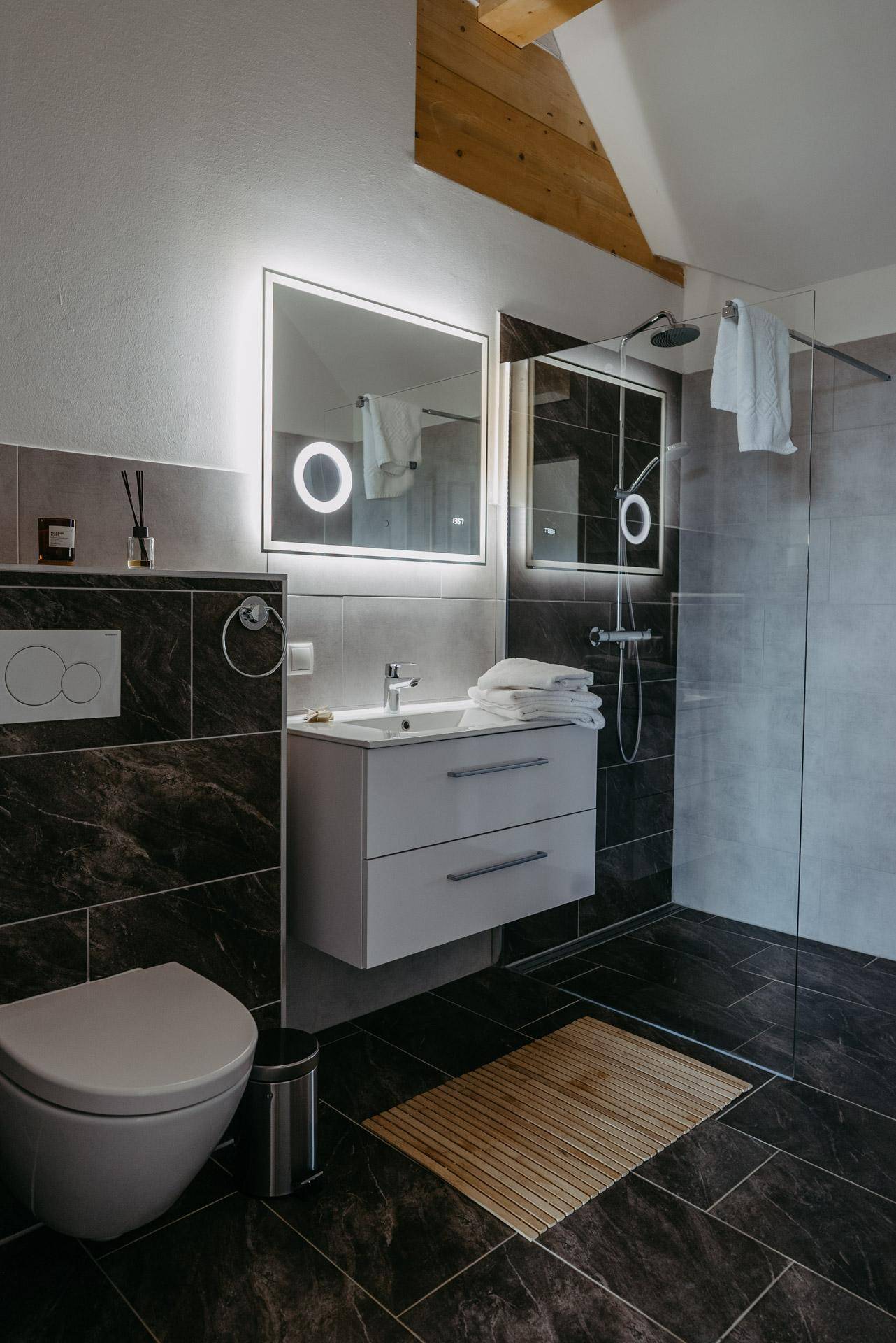 Modernes Badezimmer mit dunklen Fliesen, beleuchtetem Spiegel und ebenerdiger Dusche.