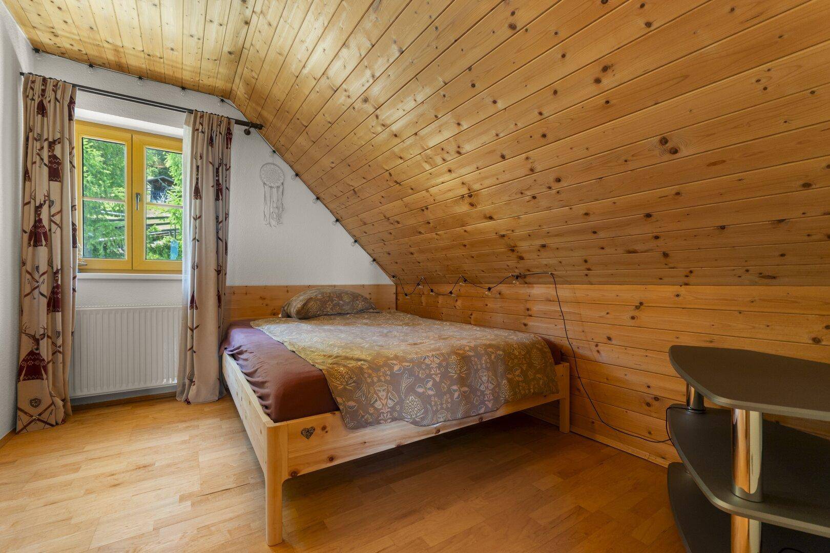 Gemütliches Schlafzimmer im Dachgeschoss mit Holzbett, schrägen Holzdecken und einem Fenster mit Blick ins Grüne.