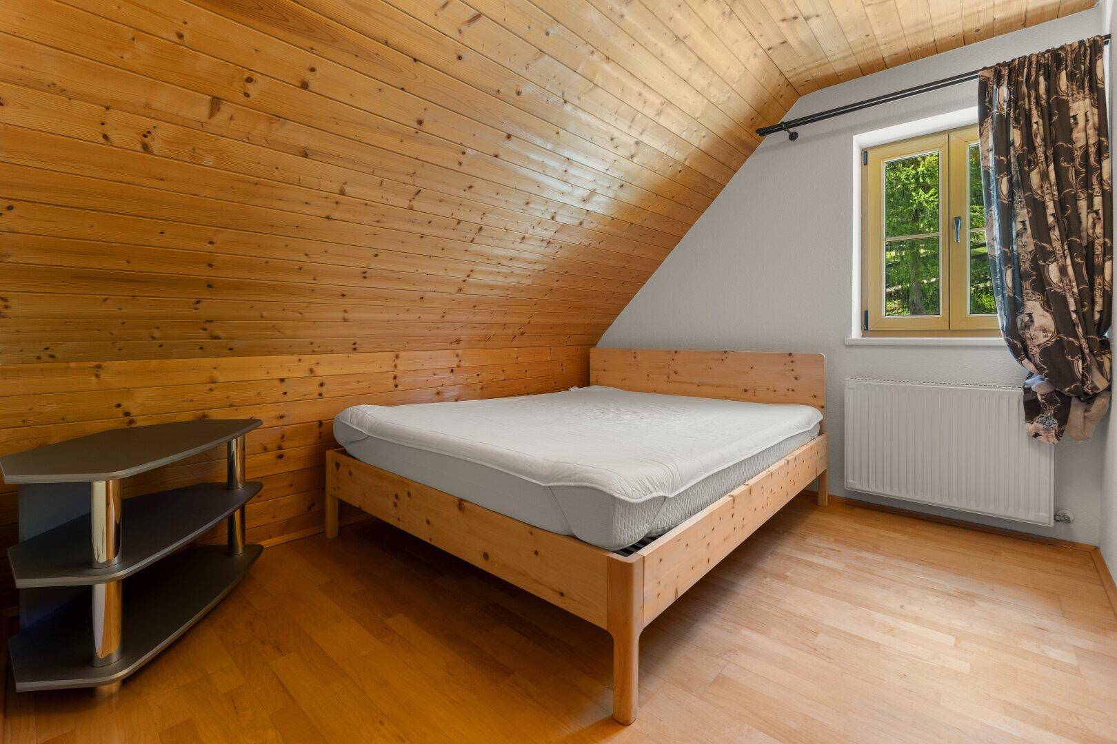 Helles Schlafzimmer im Dachgeschoss mit Holzbett, schrägen Holzdecken und einem Fenster für natürliches Licht.