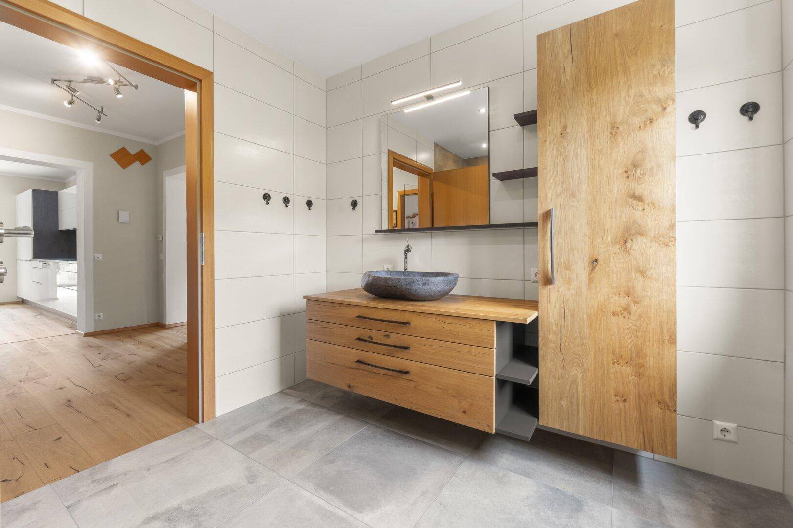 Modernes Badezimmer mit Holzelementen, Steinswaschbecken und hellen Fliesen, mit Zugang zu weiteren Räumen.