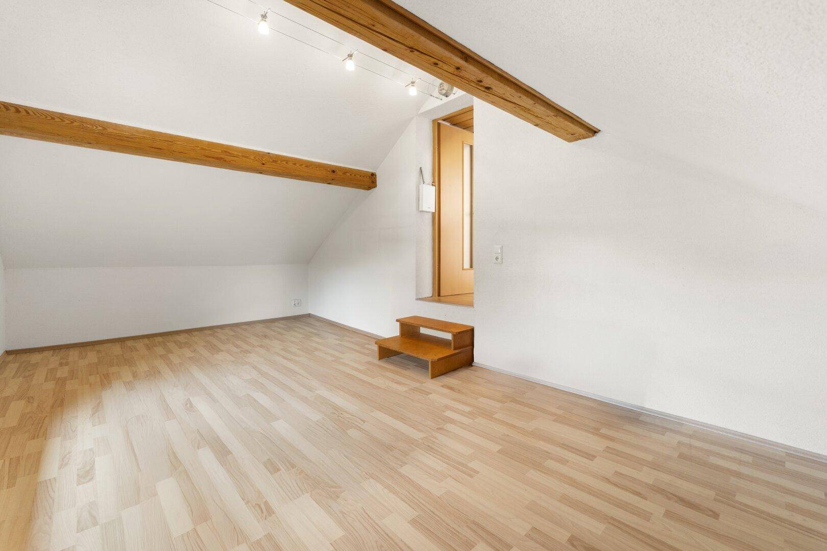 Helles Dachzimmer mit sichtbaren Holzbalken und schrägen Wänden, ausgestattet mit Laminatboden.