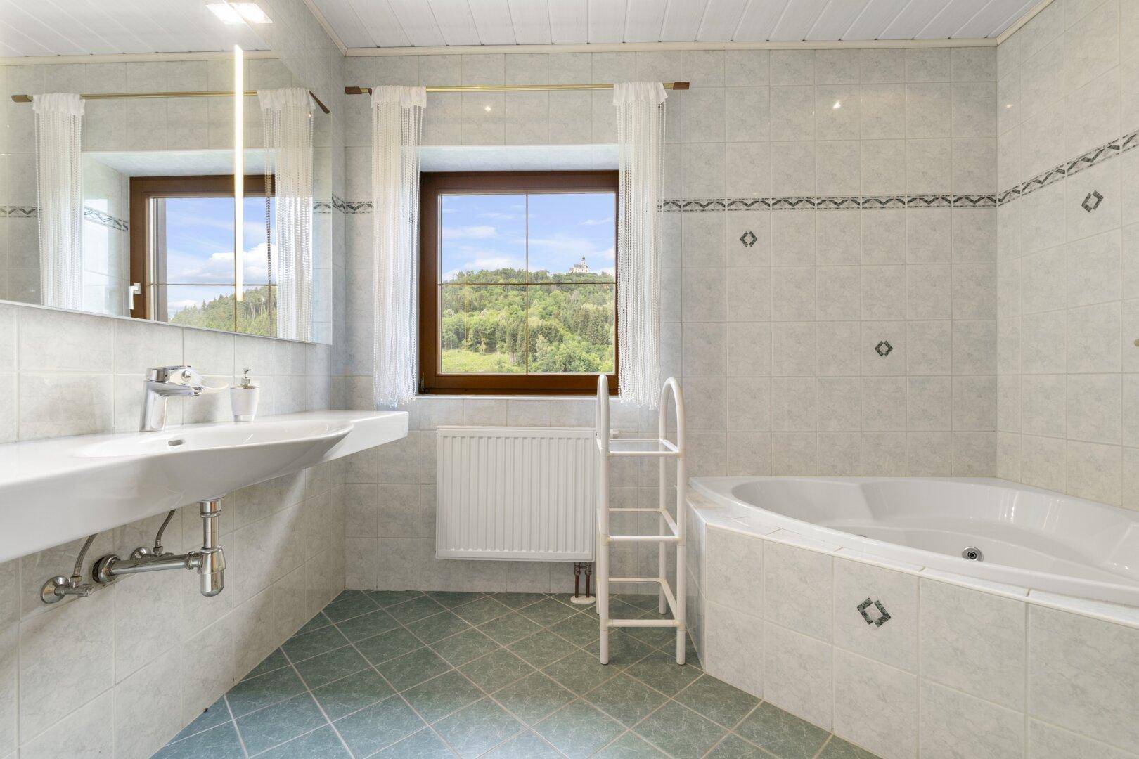 Helles Badezimmer mit hellgrauen Fliesen, Eckbadewanne und einem Fenster mit weitem Blick ins Grüne.
