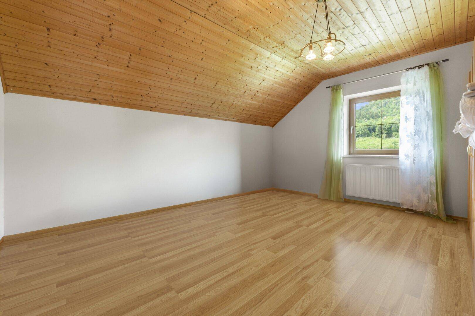 Gemütliches Dachzimmer mit Holzdecke, Laminatboden und einem Fenster mit Blick ins Grüne.