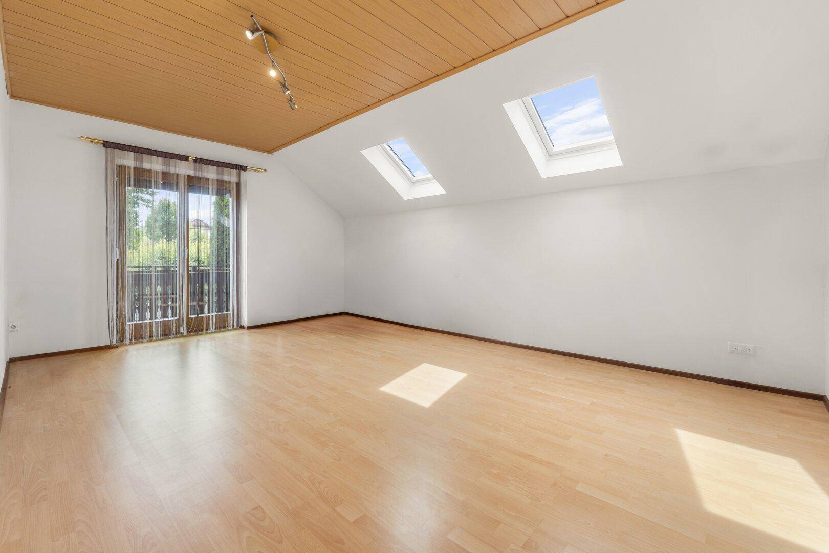 Helles Dachzimmer mit Laminatboden, Holzdecke, zwei Dachfenstern und Zugang zu einem Balkon.