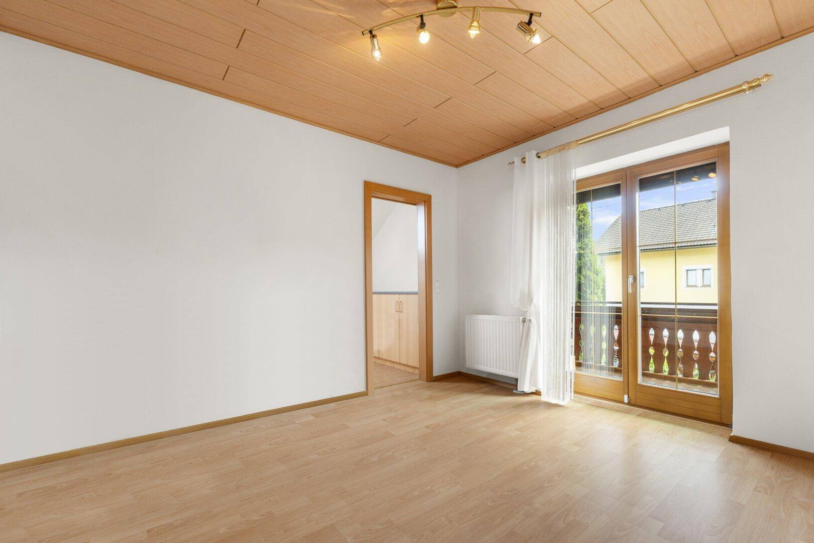 Helles Zimmer mit Laminatboden, Holzdecke und einem Balkonzugang, der viel Tageslicht hereinlässt.