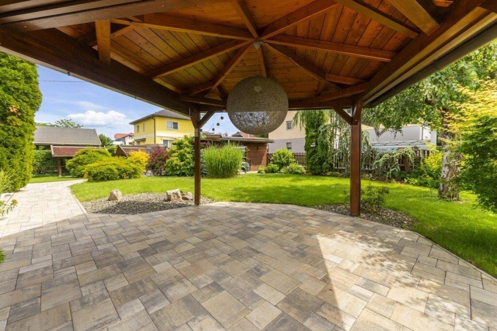 Gepflasterter Sitzbereich unter einem Holzpavillon, umgeben von einem gepflegten grünen Garten.