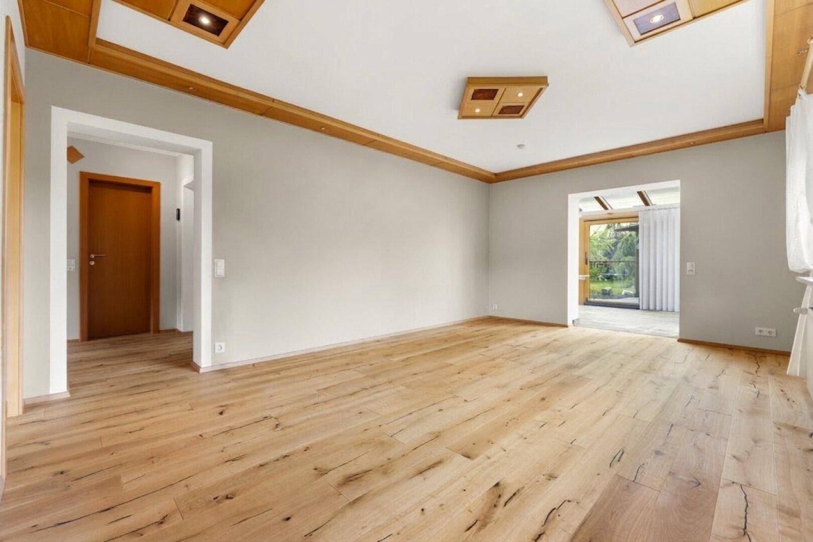 Geräumiges Wohnzimmer mit hellem Holzboden, schlichten Wänden und dekorativen Holzdeckenleisten.