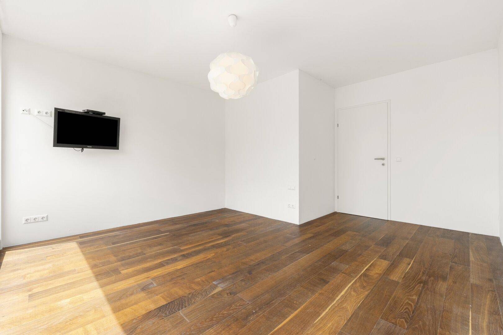 Helles Zimmer mit Holzboden und Vorbereitung für Wandfernseher, ideal als Schlaf- oder Gästezimmer.
