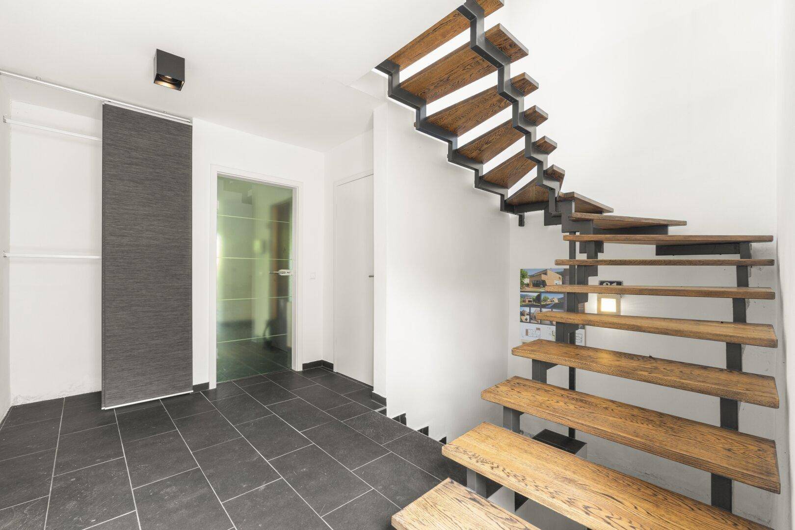 Heller Flur mit moderner Holztreppe und dunklen Fliesen, der die Stockwerke verbindet.
