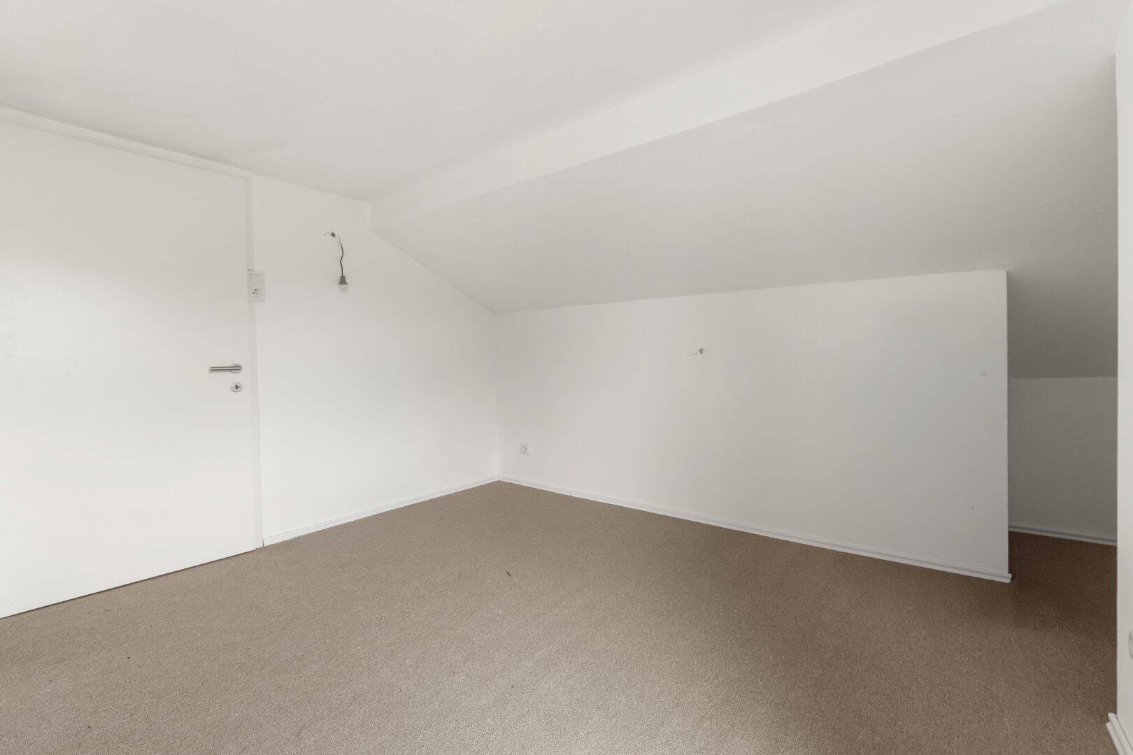 Leerer Dachbodenraum mit Teppichboden und weiß gestrichenen Wänden.