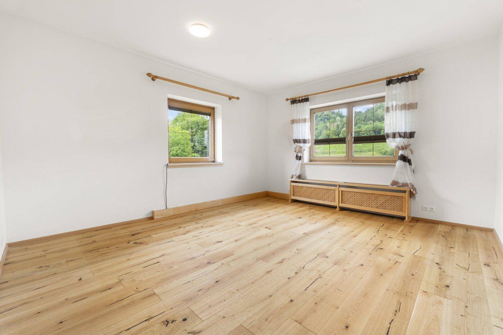 Helles Zimmer mit natürlichem Holzboden und Fenstern, die einen Blick ins Grüne bieten.