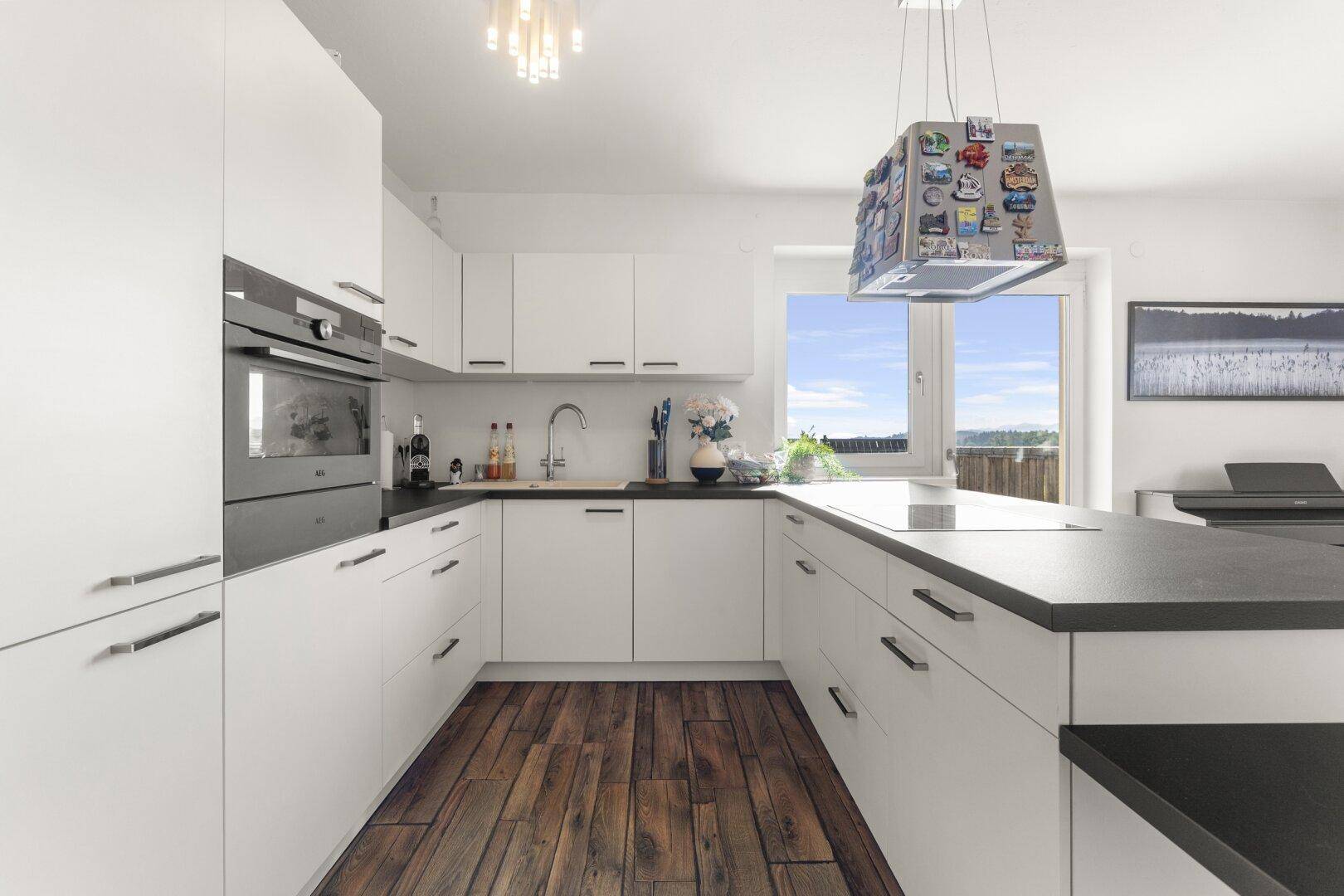 Moderne Einbauküche mit weißen Schränken, integriertem Backofen und dunklen Arbeitsplatten, mit Fensterblick.