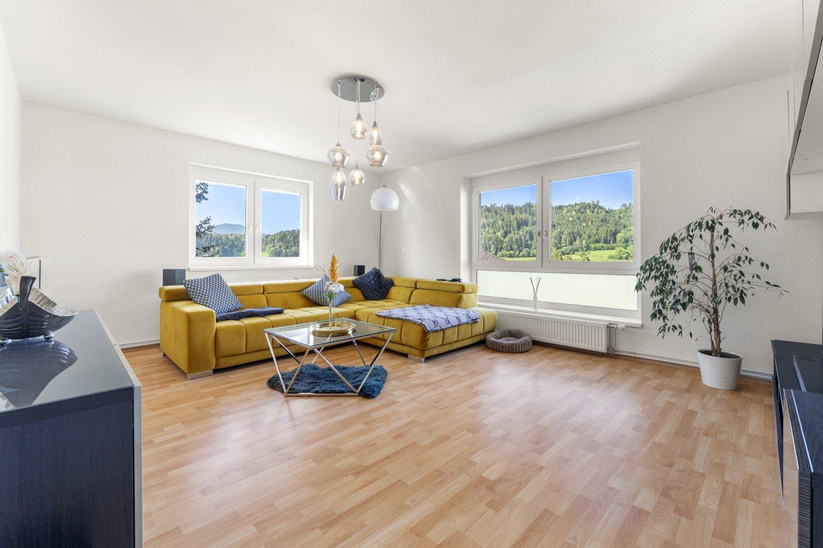 Helles Wohnzimmer mit großem gelbem Sofa und weitem Blick auf grüne Hügel und Landschaft.