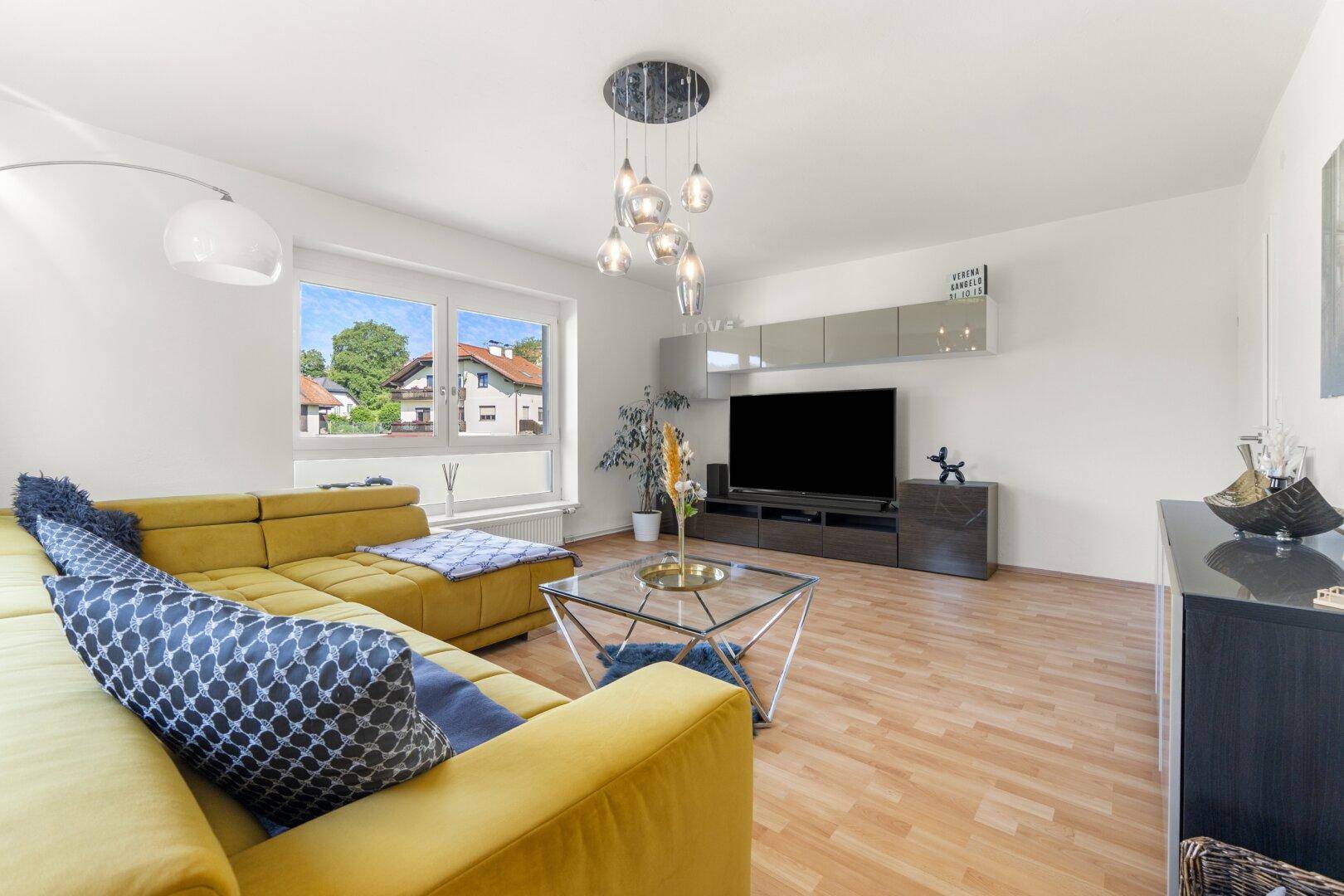 Gemütliches Wohnzimmer mit gelbem Ecksofa, Couchtisch und Fernseher, mit Blick auf die Nachbarschaft.