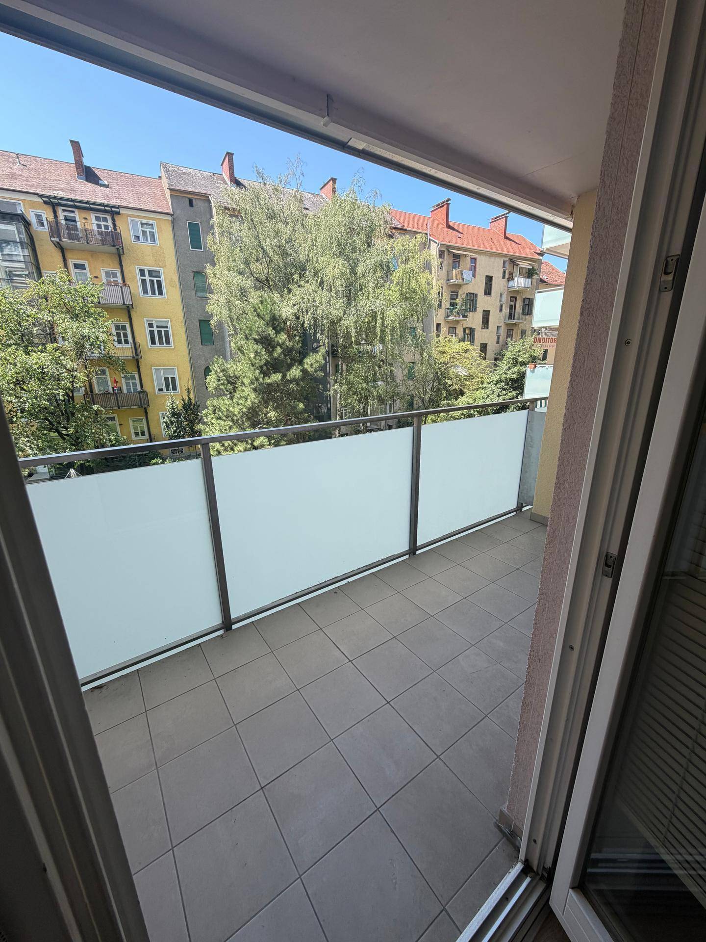Heller Balkon mit Fliesenboden und Milchglasbrüstung, bietet einen Blick auf die umliegenden Gebäude und Bäume.
