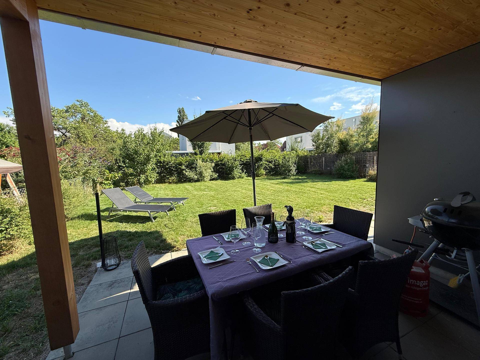 Überdachte Terrasse mit Esstisch, Sonnenschirm und Blick auf den grünen Garten an einem sonnigen Tag.