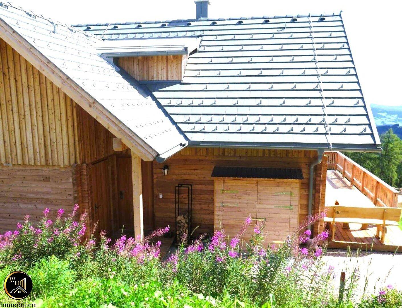 Seitenansicht des Holzhauses mit Holzlager und blühenden Pflanzen im Vordergrund.
