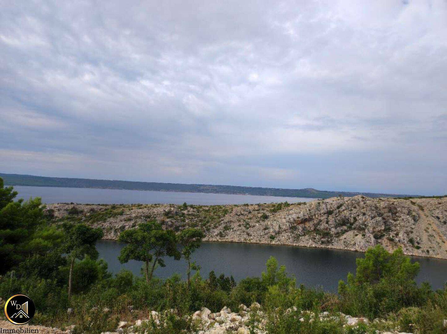 Weitläufiger Blick auf die Bucht mit felsiger Küste und grünen Bäumen unter bewölktem Himmel.