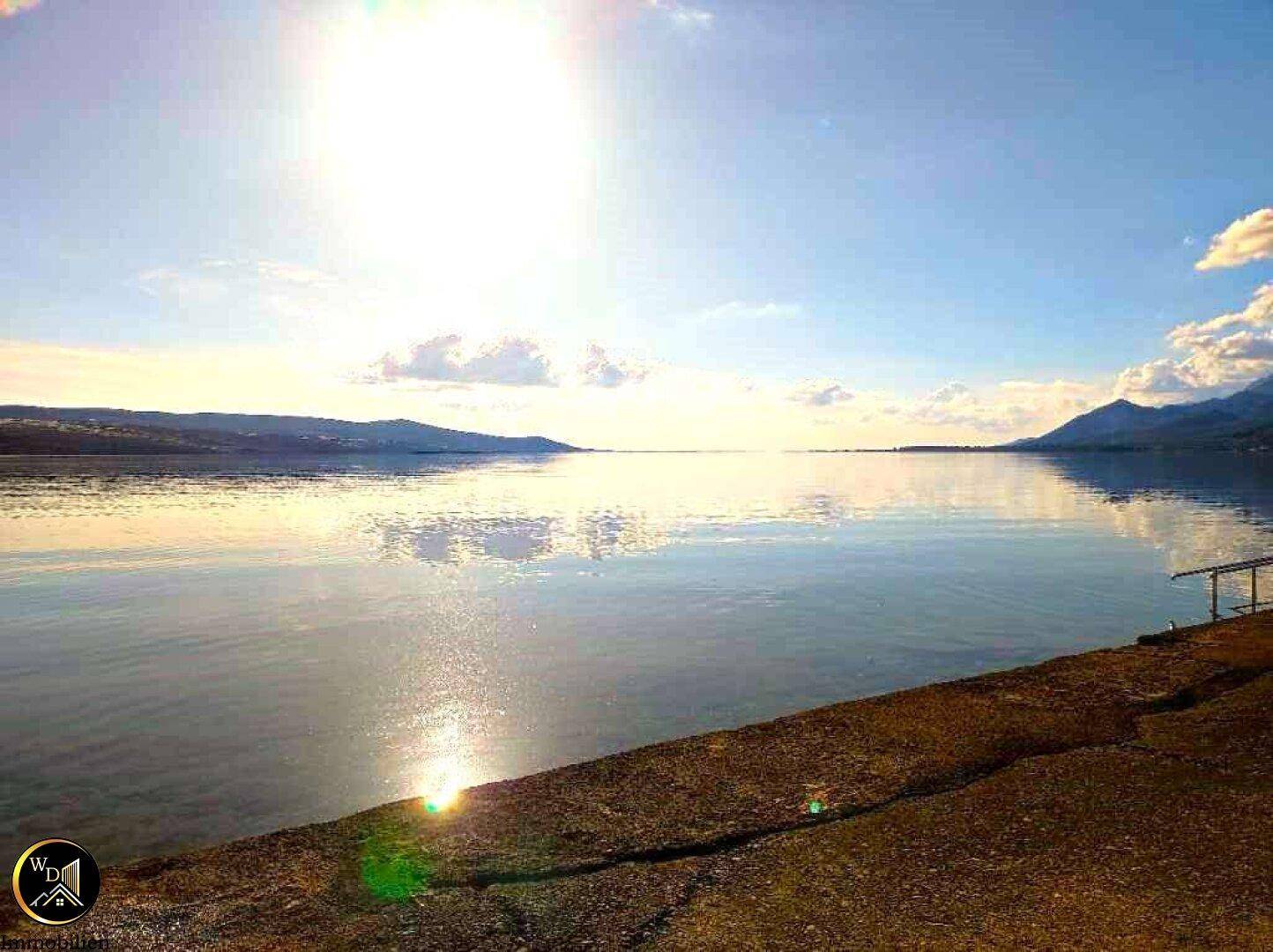 Sonnenuntergang über dem ruhigen Meer mit glitzernder Wasseroberfläche und einem Uferbereich im Vordergrund.