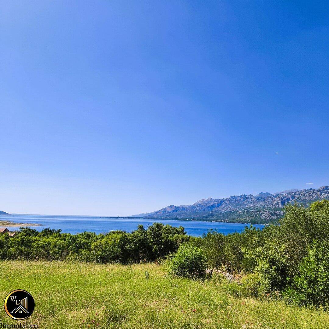 Panoramablick über grüne Hügel zum weiten Meer und den fernen Bergen unter strahlend blauem Himmel.