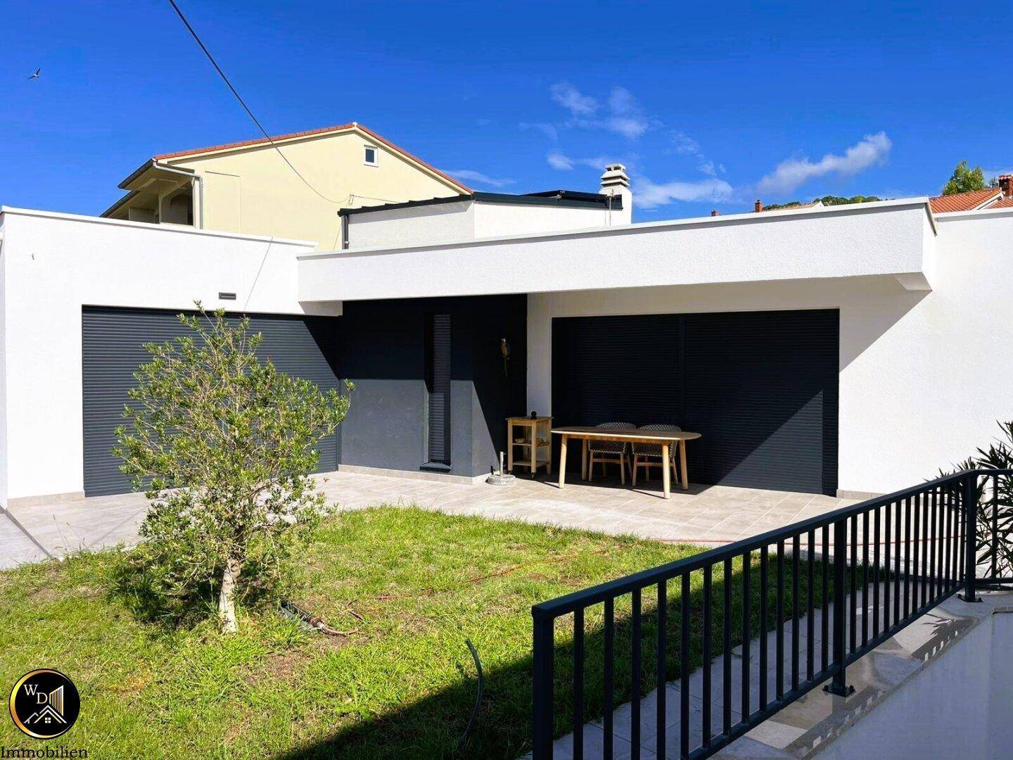 Moderne Hausfassade mit kleiner Rasenfläche und einer Terrasse mit Esstisch im Freien.