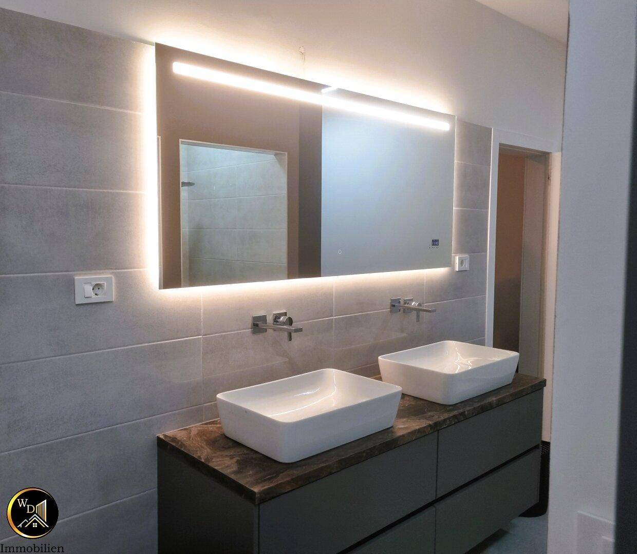 Modernes Badezimmer mit Doppelwaschbecken, beleuchtetem Spiegel und grauen Wandfliesen.