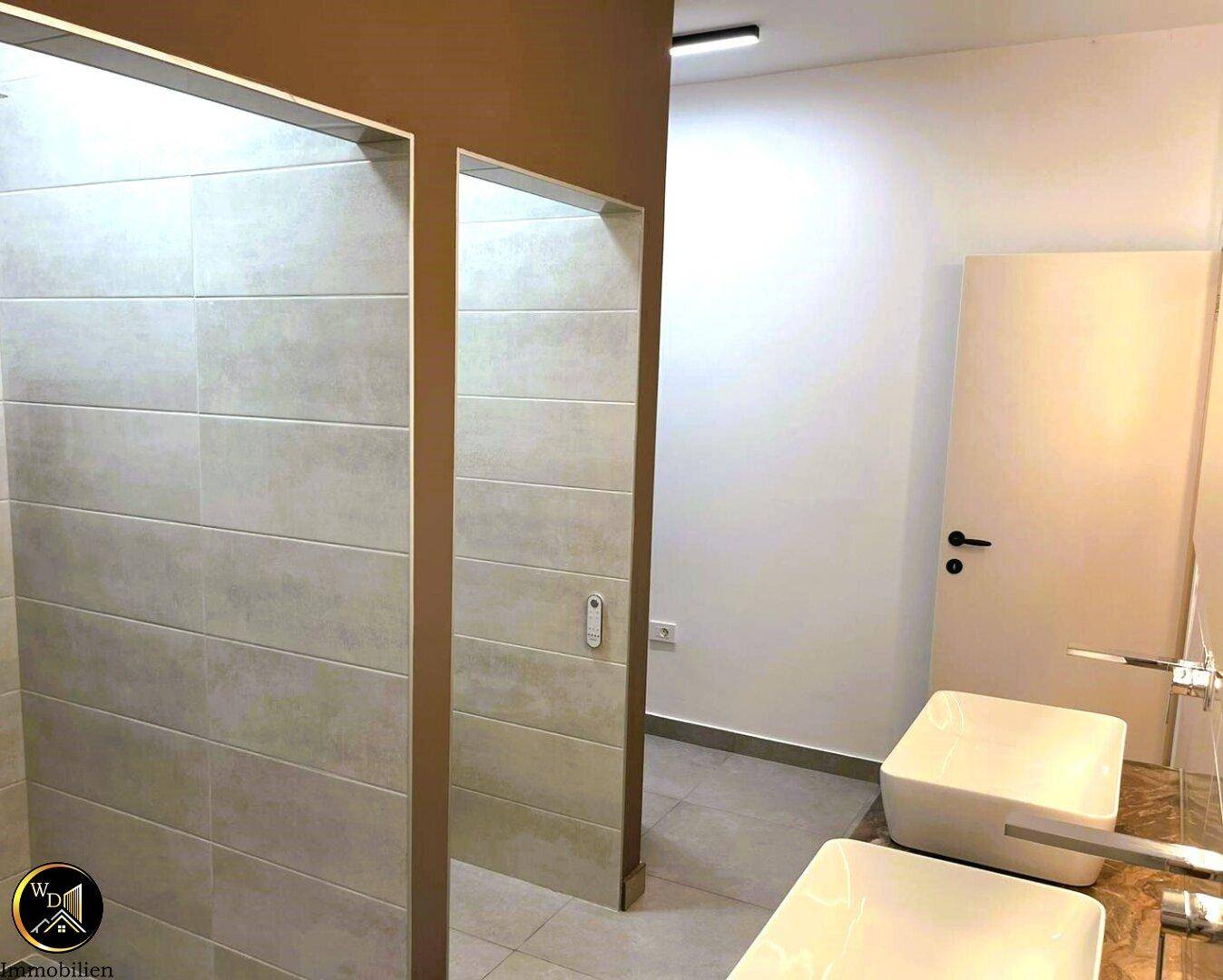 Modernes Badezimmer mit zwei Waschbecken und separaten Duschbereichen, hell gefliest.