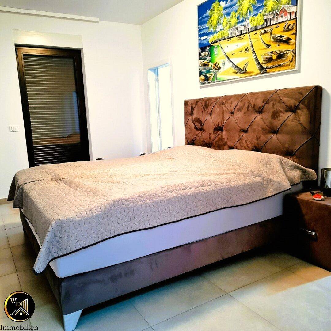 Schlafzimmer mit großem Doppelbett, gepolstertem Kopfteil und Fenster mit Rollladen.