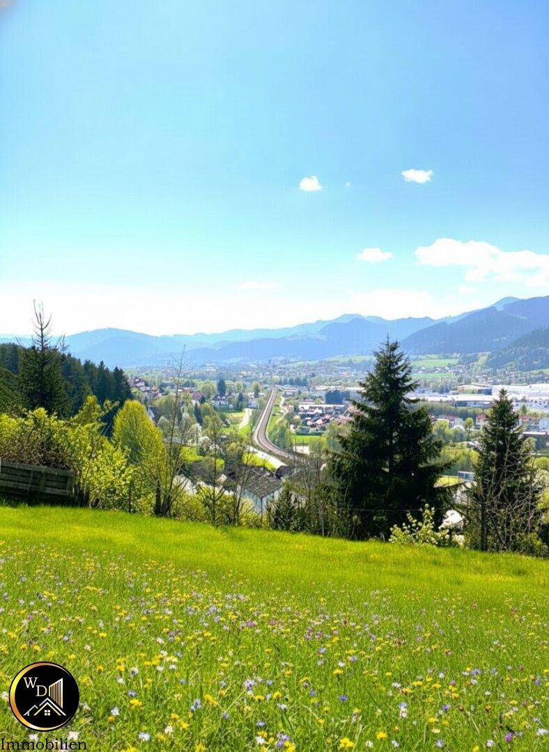 Panoramablick über ein grünes Tal mit einer Stadt, Verkehrswegen und majestätischen Bergen am Horizont.