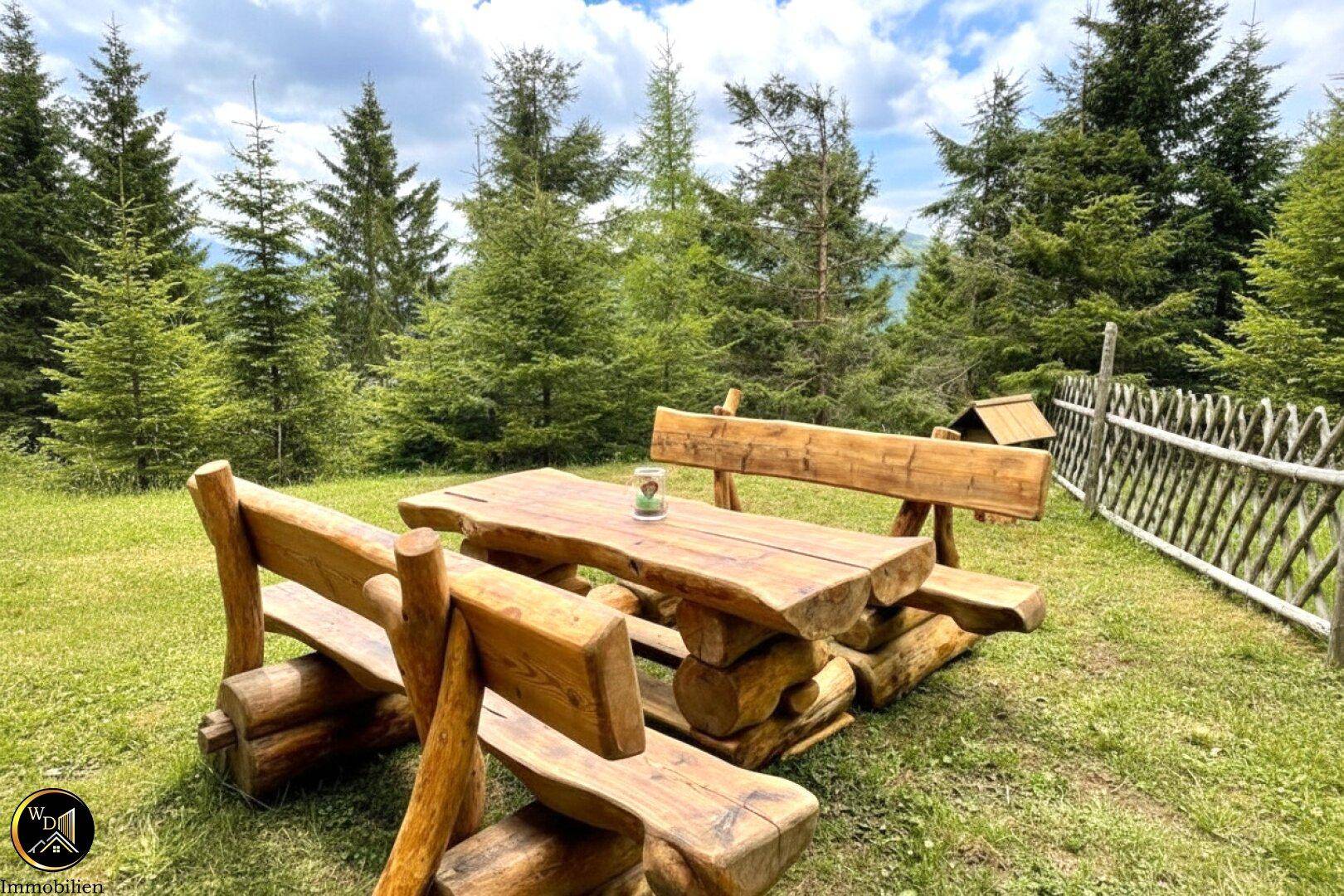 Rustikale Holzbankgruppe im Garten, umgeben von grünen Bäumen und Wiesen, ideal für Mahlzeiten im Freien.
