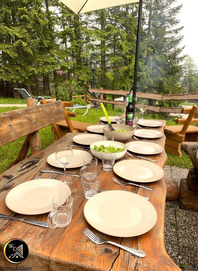 Gedeckter Holztisch im Garten, bereit für ein Essen im Freien, umgeben von grüner Natur.