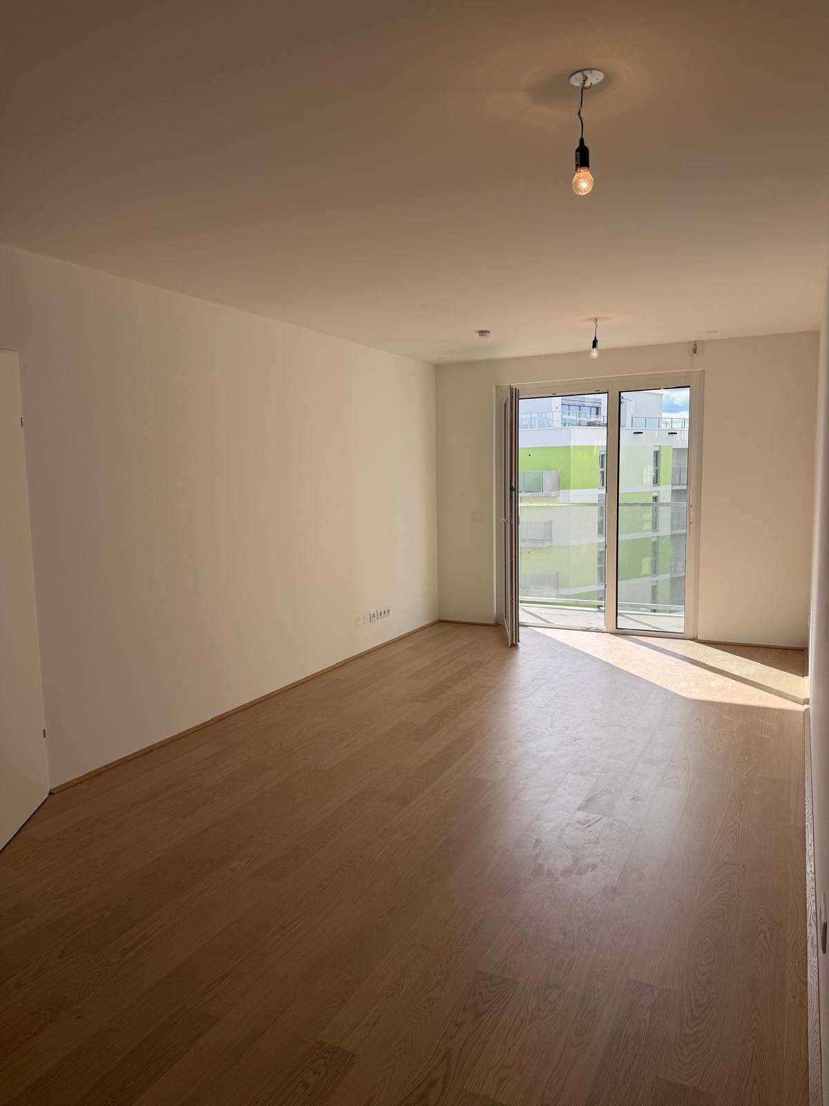 Geräumiges, helles Zimmer mit Holzboden und großem Fenster, das viel Tageslicht hereinlässt und einen Balkonzugang bietet.