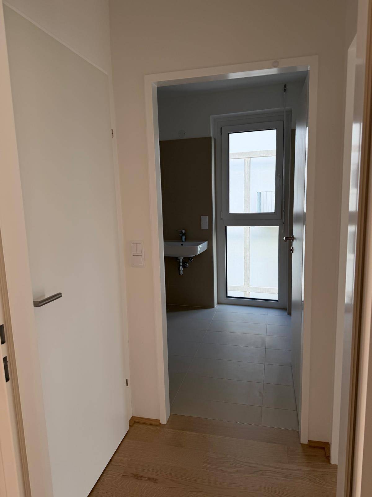 Heller Flur mit Holzboden, der zu einem Badezimmer mit Fenster und Waschbecken führt.