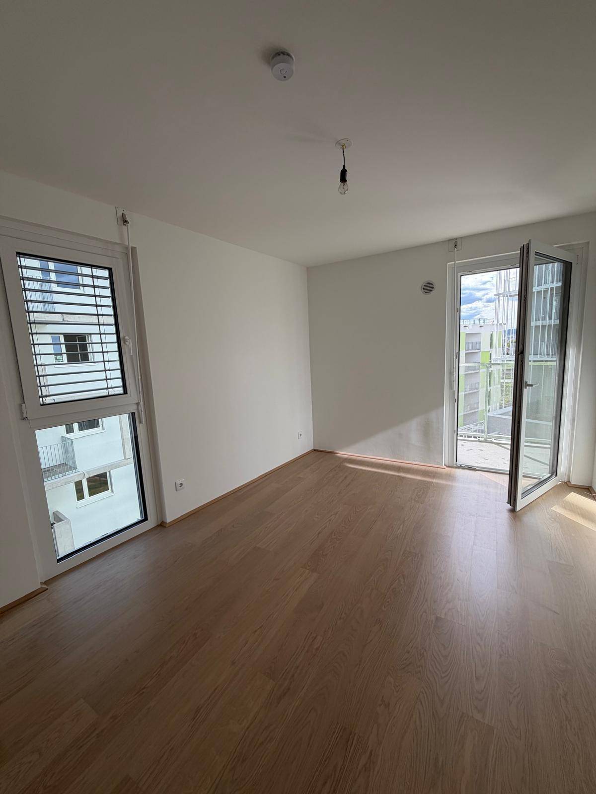 Geräumiges Schlafzimmer mit Holzboden, weißen Wänden und Fenstern, eines davon mit Außenjalousien.