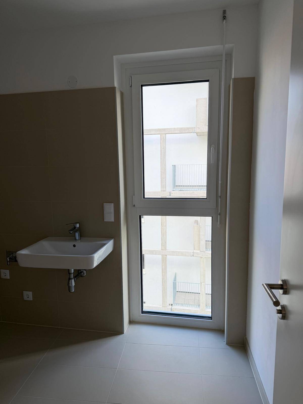 Modernes Badezimmer mit Waschbecken, hohem Fenster und beigefarbenen Fliesen, das viel Licht hereinlässt.
