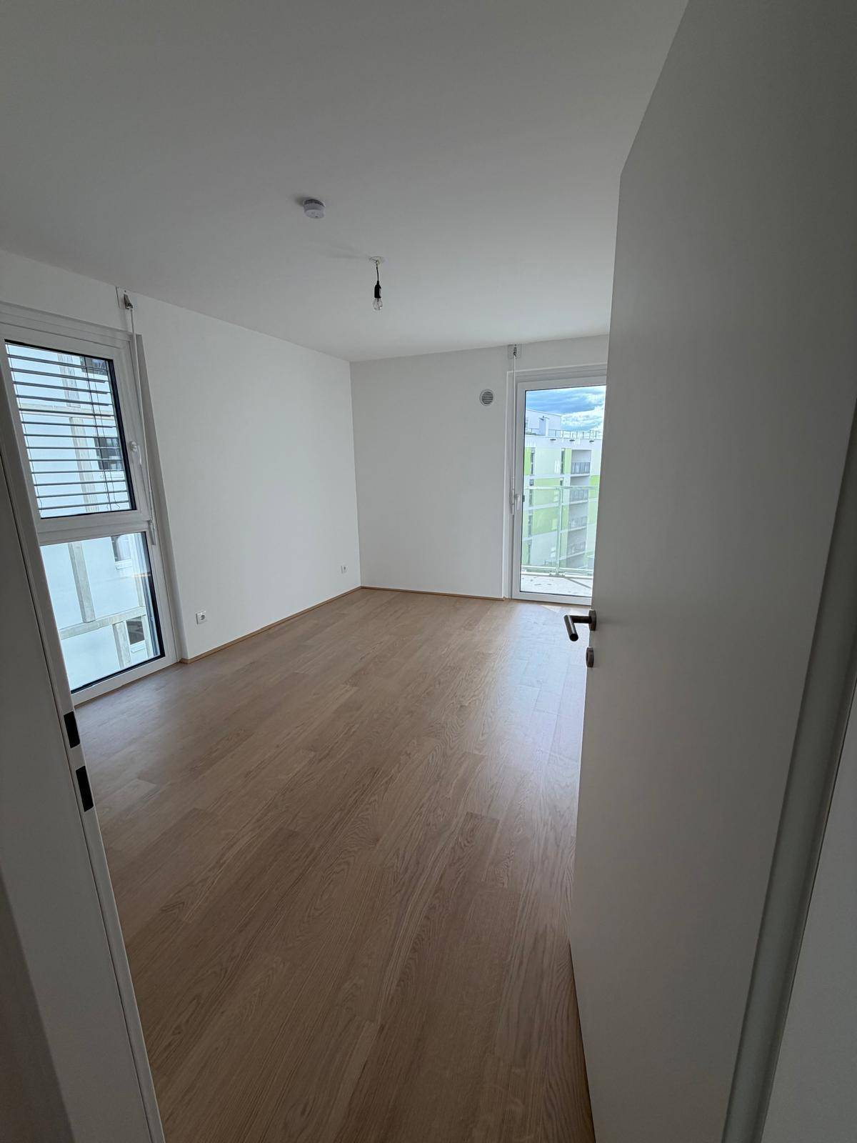 Leeres Schlafzimmer mit hellem Holzboden und zwei Fenstern, die für viel natürliches Licht sorgen.
